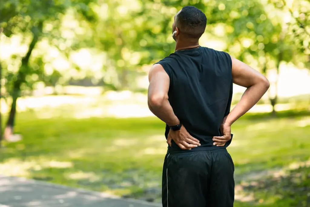 Man standing in green public area wearing workout gear and holding his lower back